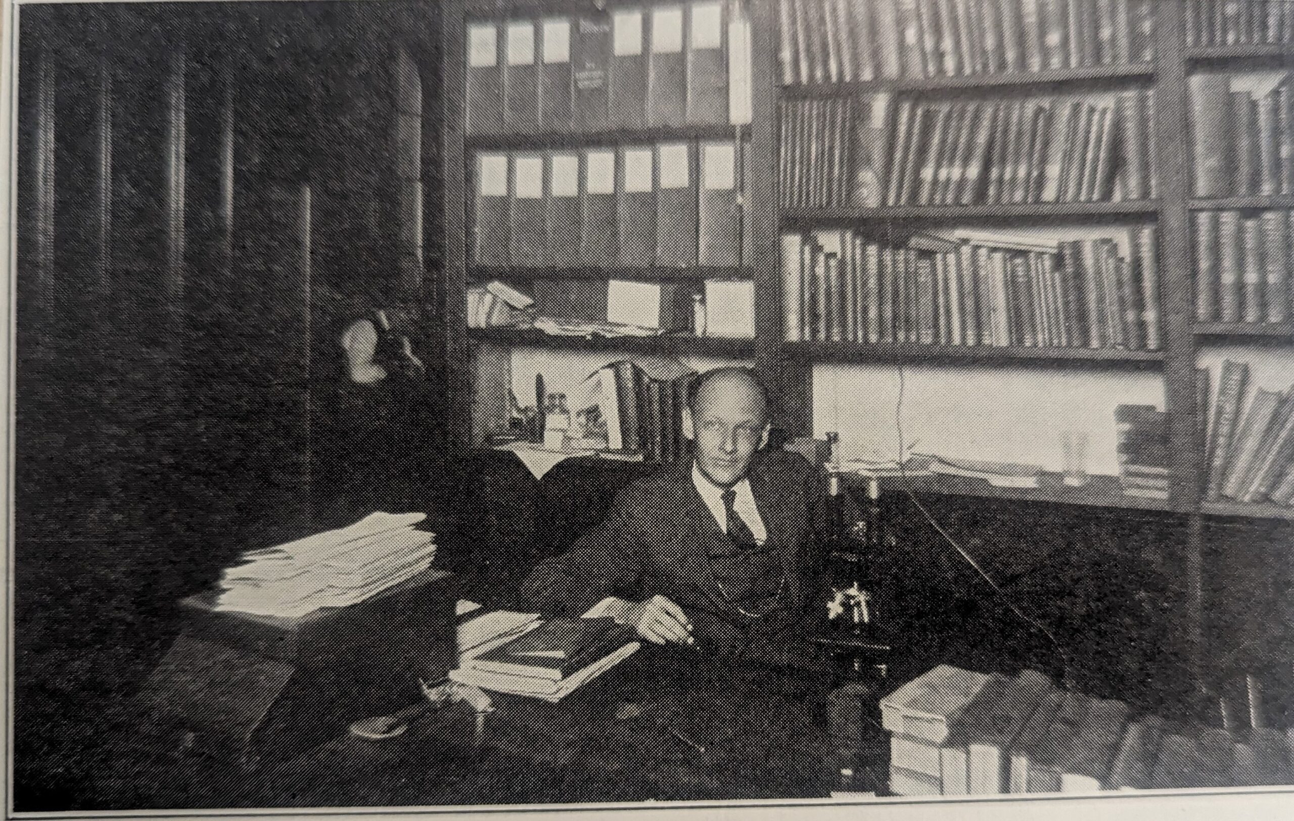 A black-and-white photograph of a man sitting at a desk in an office filled with books and documents. The man, dressed in a suit and tie, is seated with a microscope in front of him and papers spread out on the desk. Behind him are shelves filled with books and binders, suggesting a scholarly or academic setting. The lighting is dim, adding a slightly shadowed effect to the image.