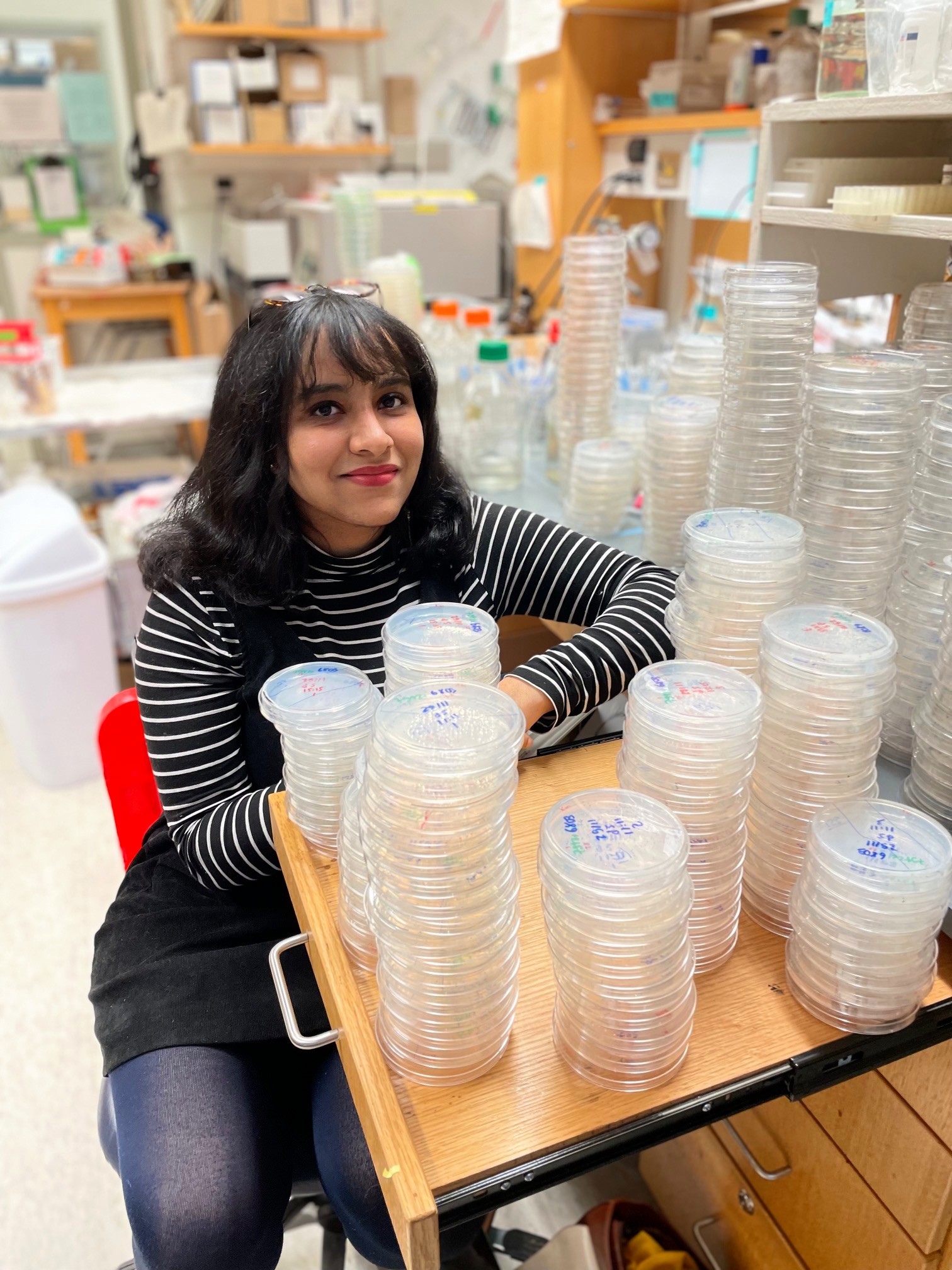 A young scientist with medium-length dark hair sits in a laboratory surrounded by stacks of clear petri dishes on a wooden table. She is wearing a black and white striped long-sleeve shirt and black dress, and she is smiling while looking at the camera. The background includes various lab equipment and supplies.