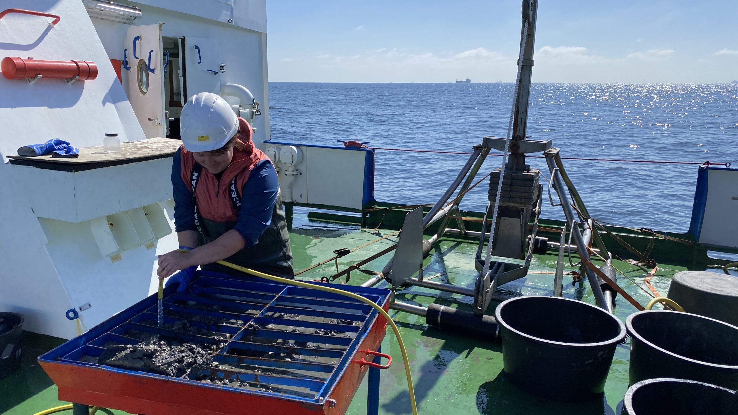 A person on the deck of a research vessel, wearing a white hard hat, a blue and red jacket, and blue gloves. They are hosing off mud from a grid-like metal structure that appears to be a sieve or sediment sampler. The sea is visible in the background under a clear sky, and there is equipment on the deck surrounding the person.