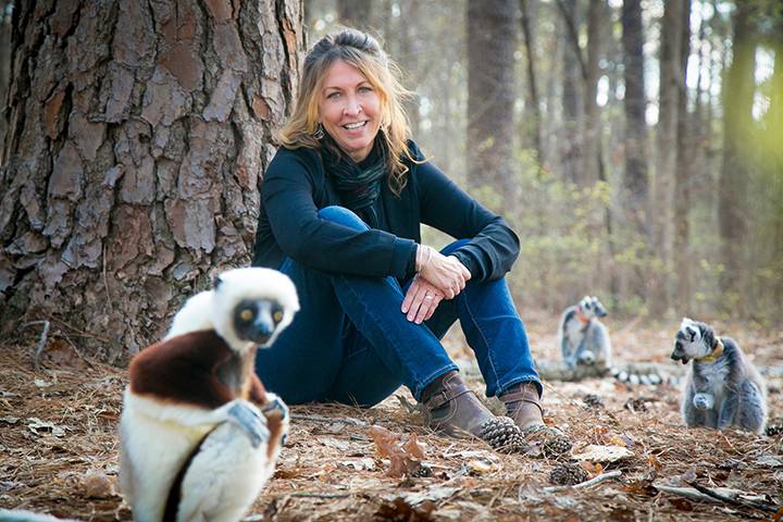 Anne Yoder sitting in front of a tree with a couple lemurs in the background and one in the foreground