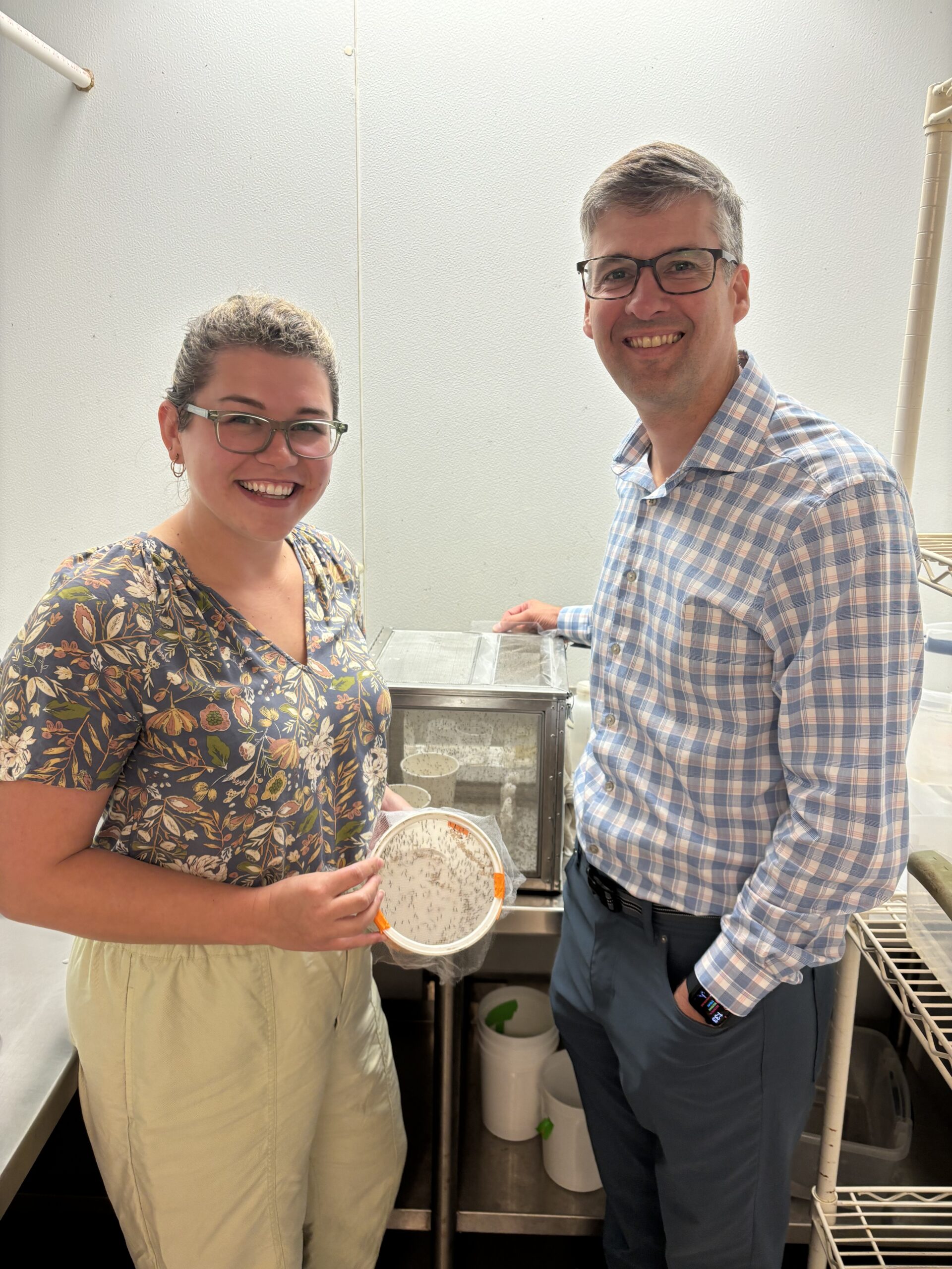 Jordyn Barr and Julian Hillyer are standing together in a laboratory setting. Jordyn, on the left, is smiling and holding a container with small organisms inside. She is wearing glasses, a floral-patterned blouse, and light-colored pants. Julian, on the right, is also smiling and wearing glasses, along with a checkered shirt and dark pants. Both are standing near lab equipment, with a clean white backdrop typical of a laboratory environment.