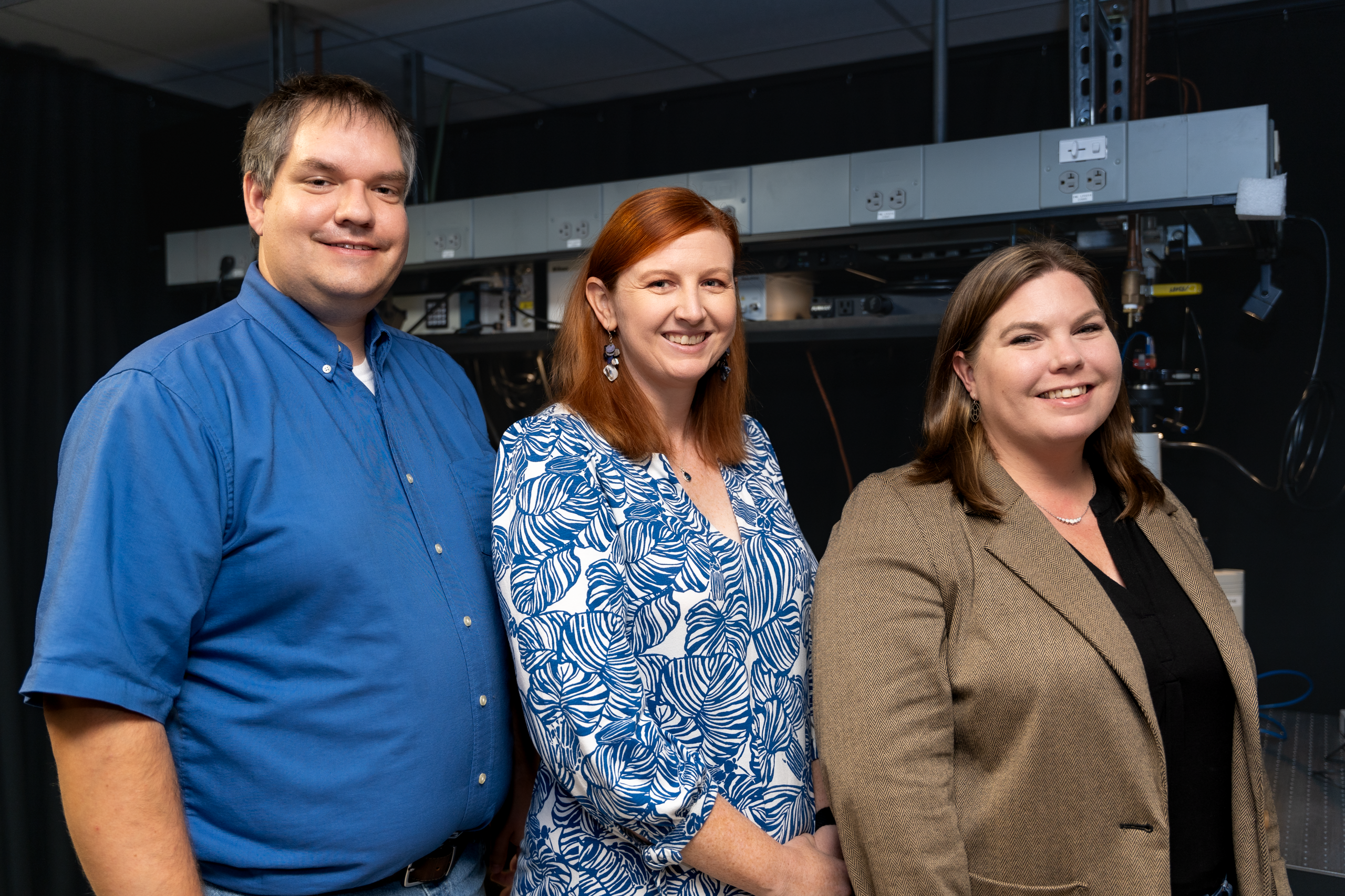 The image shows three individuals standing side by side in a professional setting. From left to right: A man wearing a blue button-up shirt, smiling at the camera. A woman with red hair wearing a blue and white patterned blouse, also smiling. A woman with brown hair wearing a beige blazer over a black top, smiling. They are standing in front of a backdrop of a laboratory environment with equipment visible in the background.