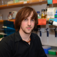 Bryan with long hair and a dark t-shirt. He is smiling sitting in a lab setting