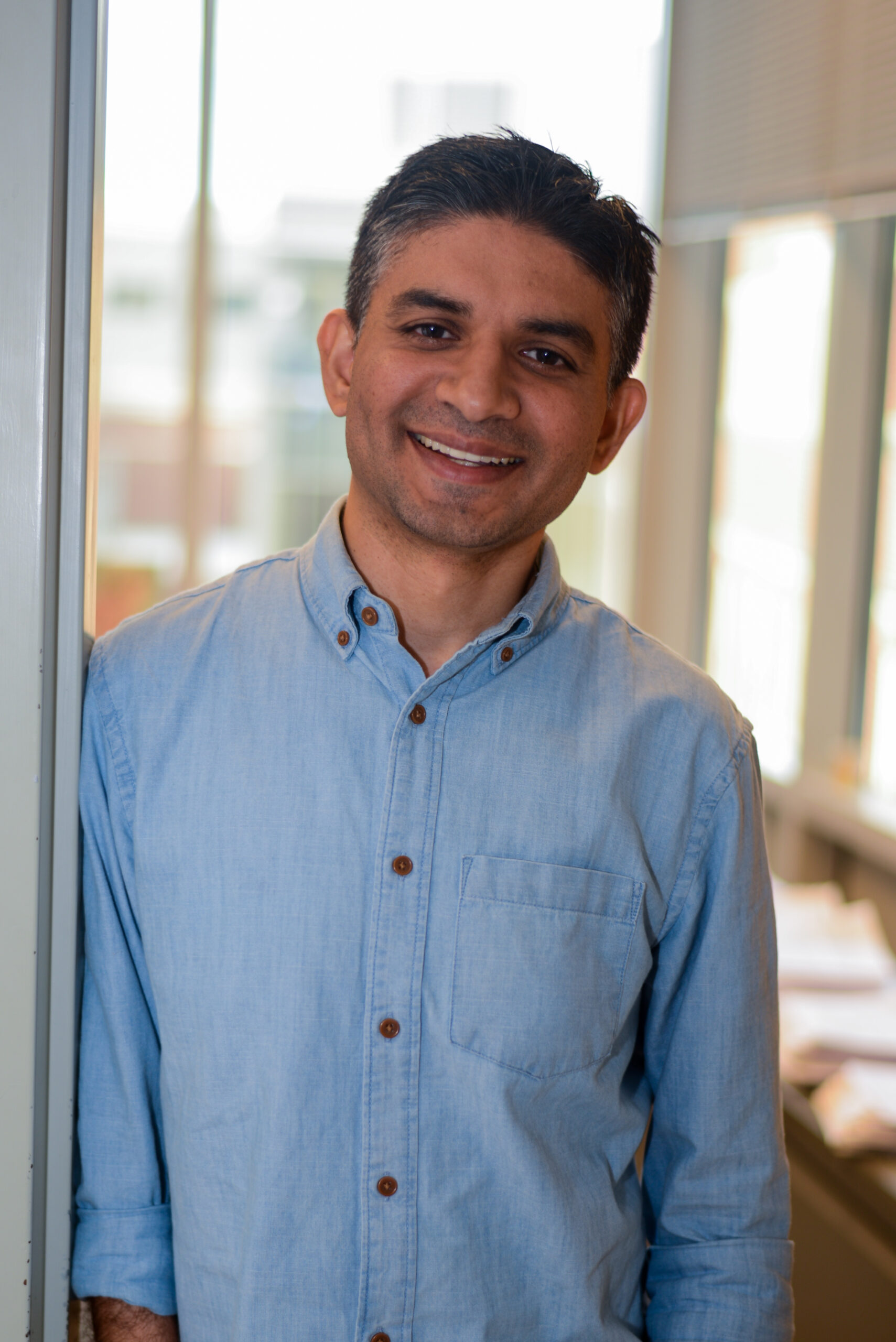 Maulik smiling in front of a lab window. He is wearing a light blue button up shirt.