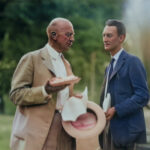 An older man with glasses and hearing aids, dressed in a tan suit, holding a piece of bread and a hat, is in conversation with a younger man in a blue suit and tie outdoors, surrounded by greenery