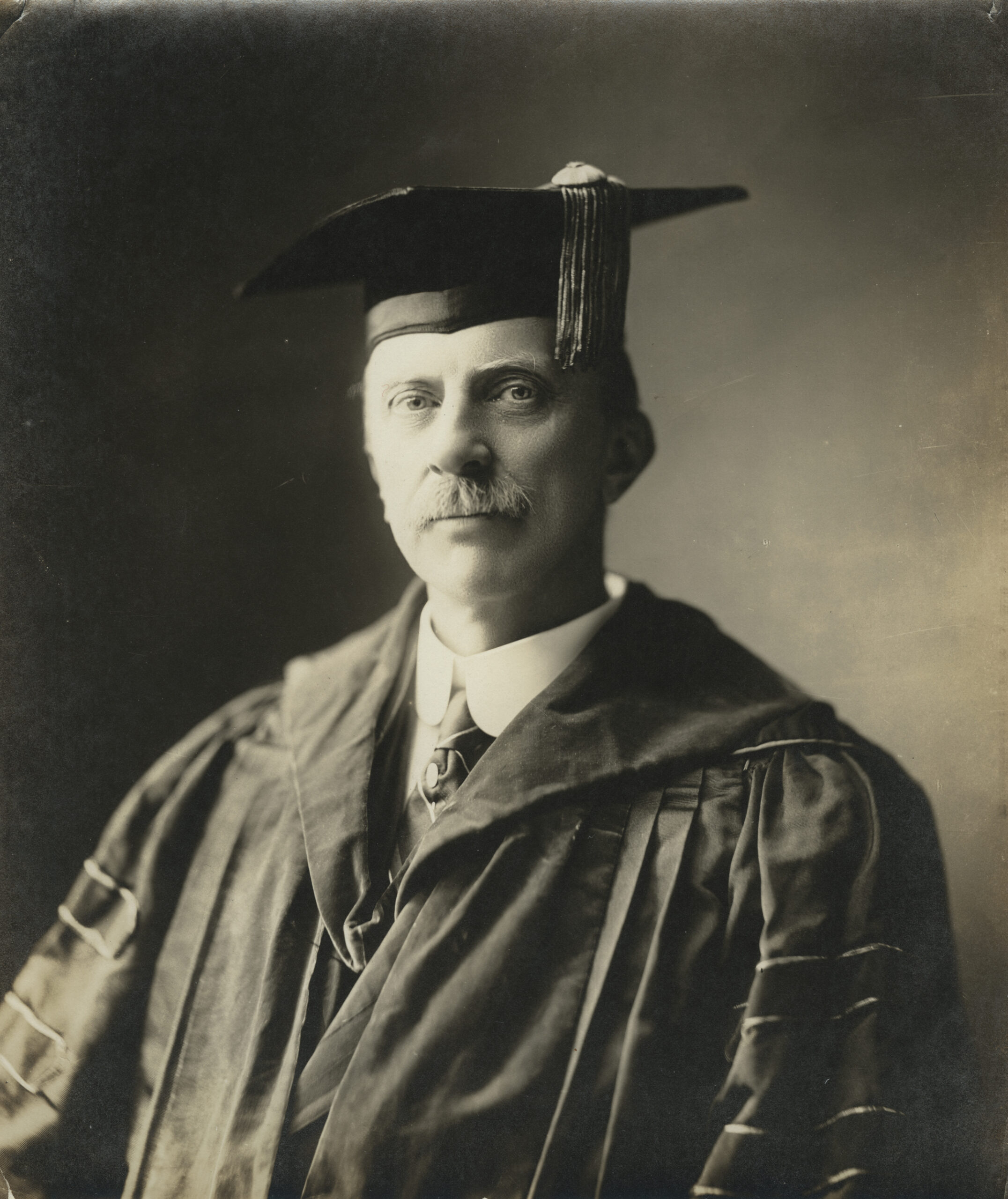 Portrait of a middle-aged man with a mustache, wearing academic regalia including a cap and gown, looking directly at the camera with a serious expression.