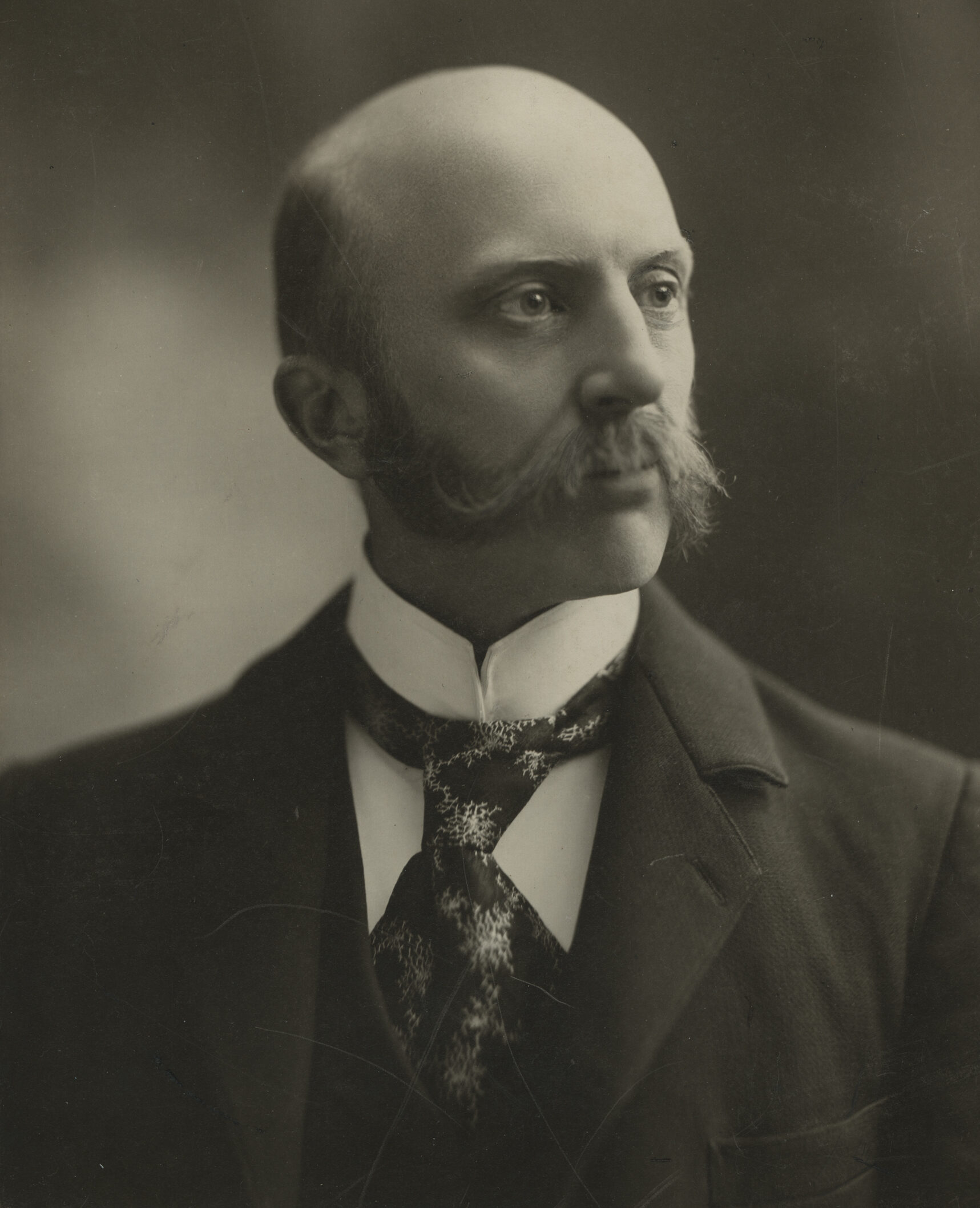 Greyscale portrait of a bald man with a distinctive handlebar mustache, dressed in formal attire with a patterned cravat, gazing off to the side in a serious manner.