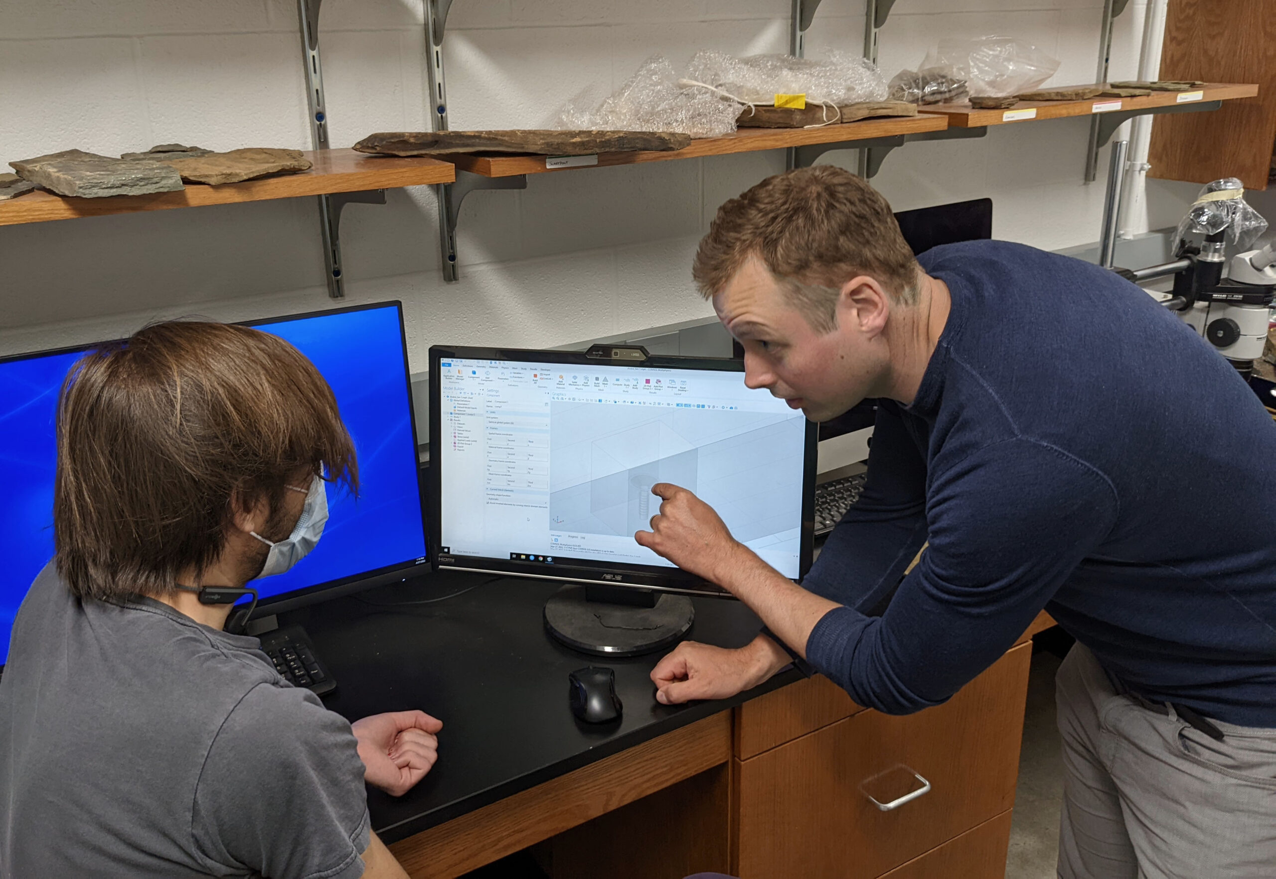 A person in a blue shirt is pointing at a computer screen displaying a 3D model, explaining something to another individual seated beside them, who is wearing a face mask and a Rolling Stones t-shirt. The room has shelves with rock samples above the desk.