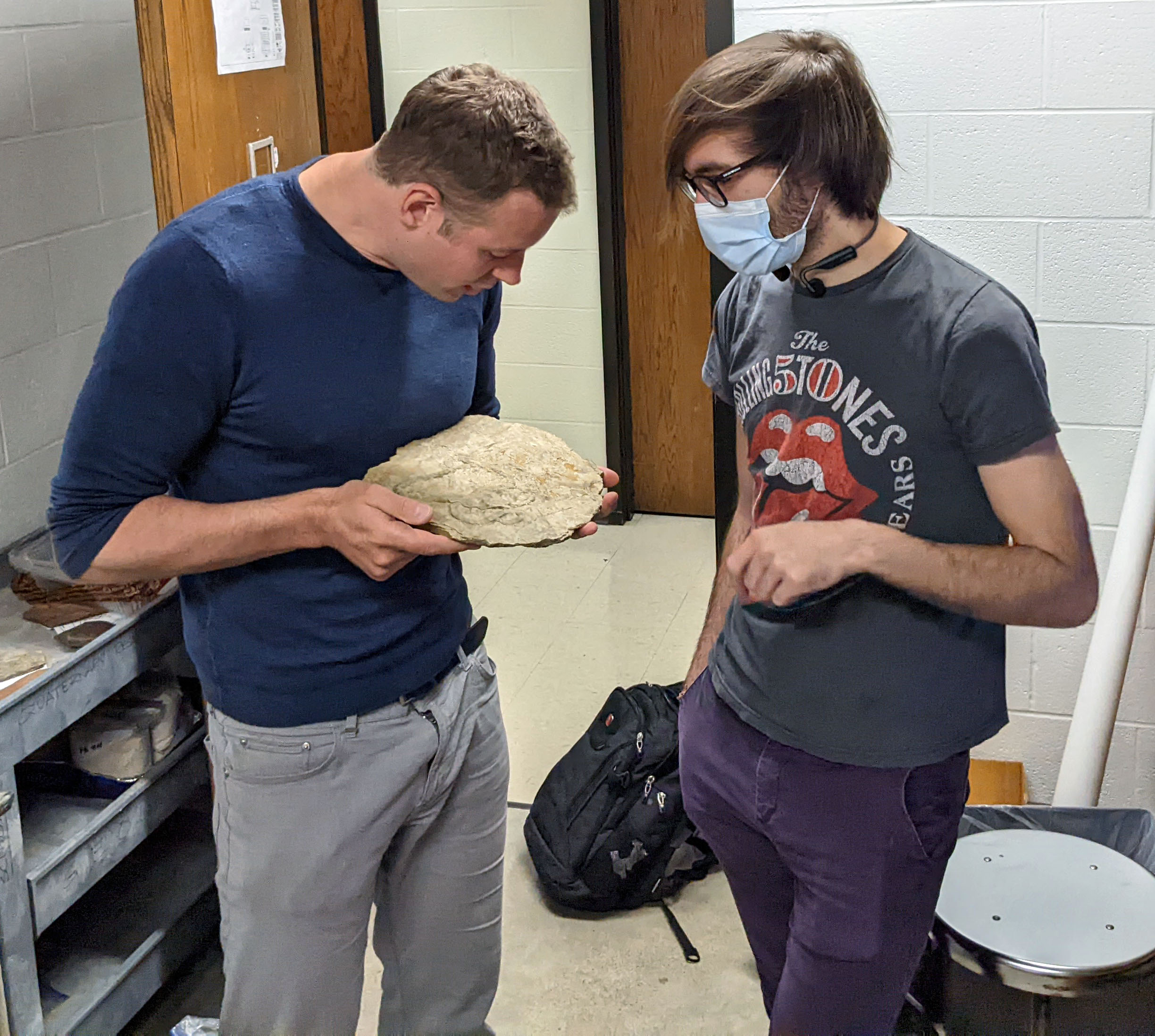 A person in a blue shirt is closely inspecting a large rock, holding it in both hands, while another individual in a Rolling Stones t-shirt and a face mask stands beside them, gesturing toward the rock. They appear to be discussing the sample in a room with additional rock samples on nearby shelves.