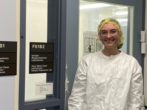 Sylvia Cheever, a Ph.D. student in Biological Anthropology, stands outside a trace metal clean laboratory in protective lab gear, including a yellow hair cover, goggles, and a white lab coat.