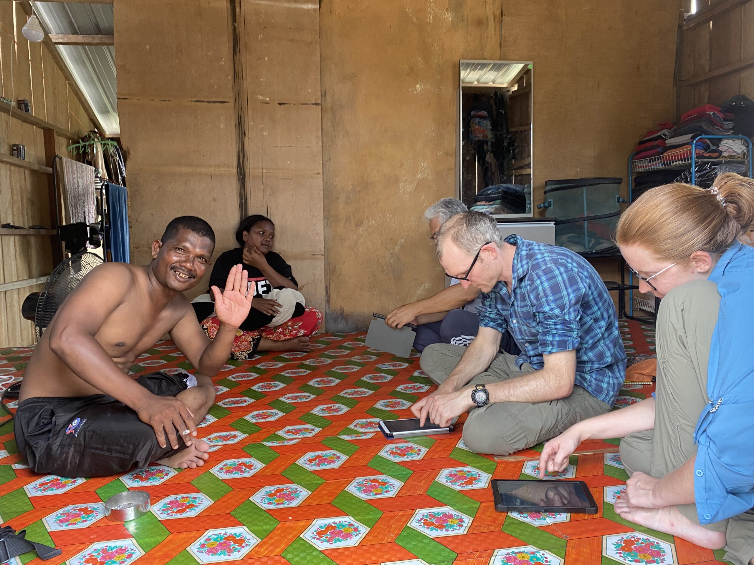 A group of people is seated on a brightly patterned mat inside a rustic room. A local man smiles and waves at the camera, while others, including two researchers using tablet devices, sit nearby. The background includes personal belongings and a small fan, giving the space a homely feel.