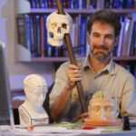 Ken Catania sitting in his office holding a staff with a skull reproduction.