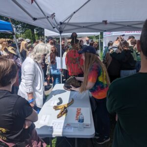 A lively outdoor event featuring a table under a white canopy. People are gathered around, interacting with items on display, including a crocodile skull, fossils, and other scientific artifacts. A person in a tie-dye shirt is explaining something to attendees. In the background, more people are exploring similar booths