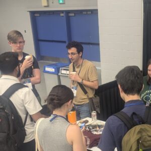 "A man in a brown shirt holding a coffee cup is engaged in conversation with a small group of people at an indoor event. He is wearing a lanyard and is part of an animated discussion, with a blue door and a booth table partially visible nearby.