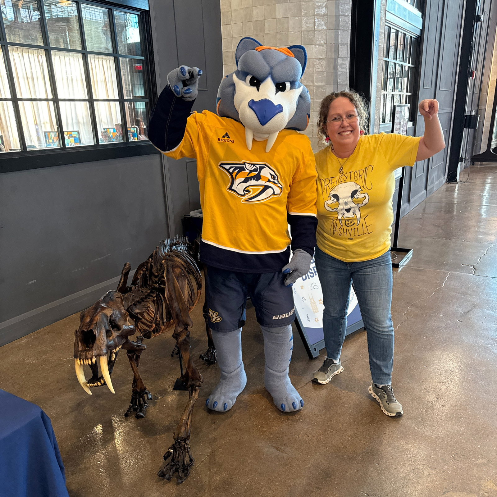 Larisa DeSantis and Gnash, the Nashville Predators mascot, pose playfully with raised fists next to a saber-toothed cat skeleton. Larisa wears a yellow "Prehistoric Nashville" shirt, and Gnash sports a Predators jersey. They stand in a brightly lit indoor space.
