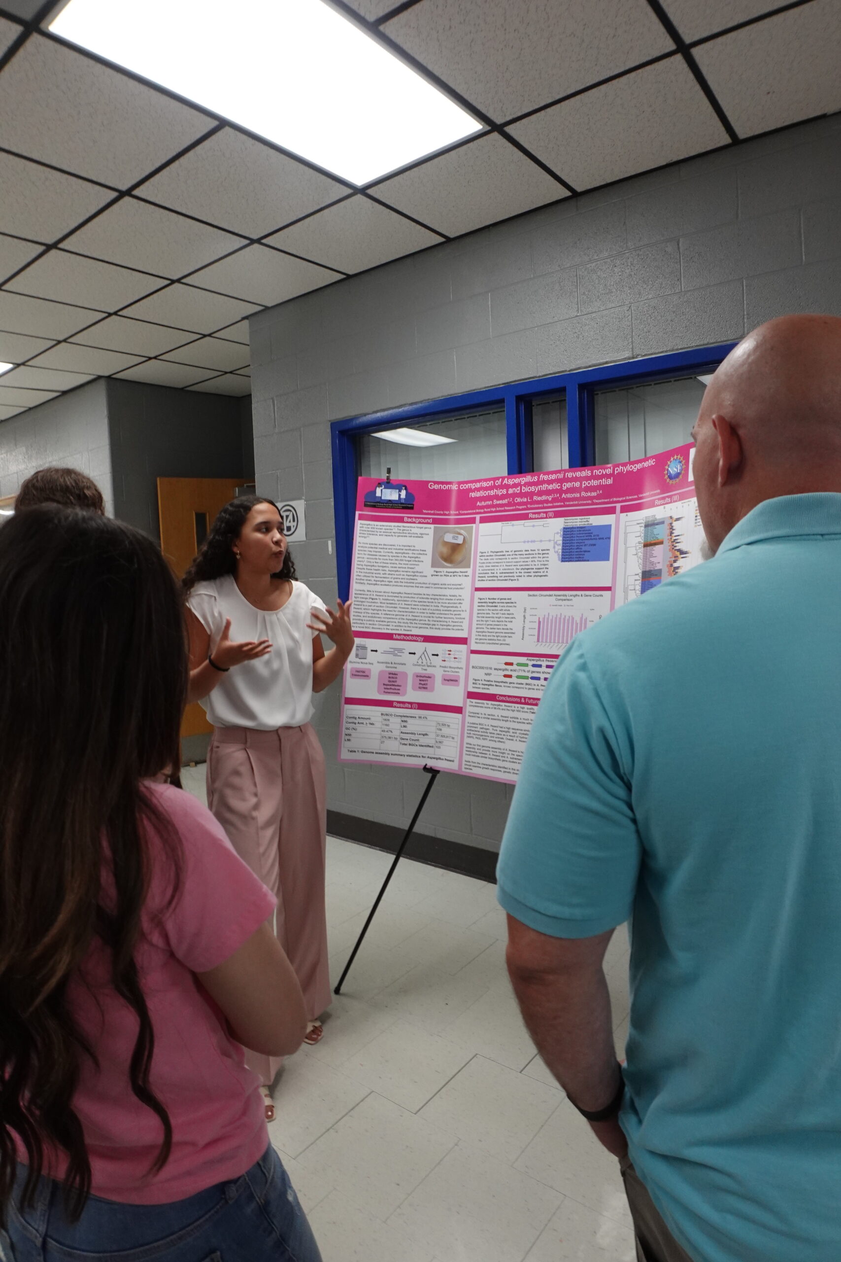 A young woman presents a bright pink scientific poster titled "Genomic comparison of Aspergillus..." to a small group of adults in a hallway with gray brick walls and fluorescent lighting. She gestures animatedly while explaining her work. The group listens attentively, their backs partially turned to the camera.