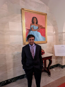 Image is portrait and shows the new portrait of Michelle Obama, hanging in the White House, wearing a blue ballgown with a pink background, she sits, with her left elbow draped onto the side of the chair. Below the portrait, slightly obscuring it's bottom left side, stands Hari, in a black suit with a purple shirt and tie. 
