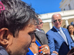 Image shows a close up of Hari shaking hands with President Biden. Hari's face can be seen to the left of the image focused on his right. President Biden, sporting his often-seen Aviator sunglasses and a blue suit, is to the right hand side of the frame, slightly out of focus, and looking toward Hari. 