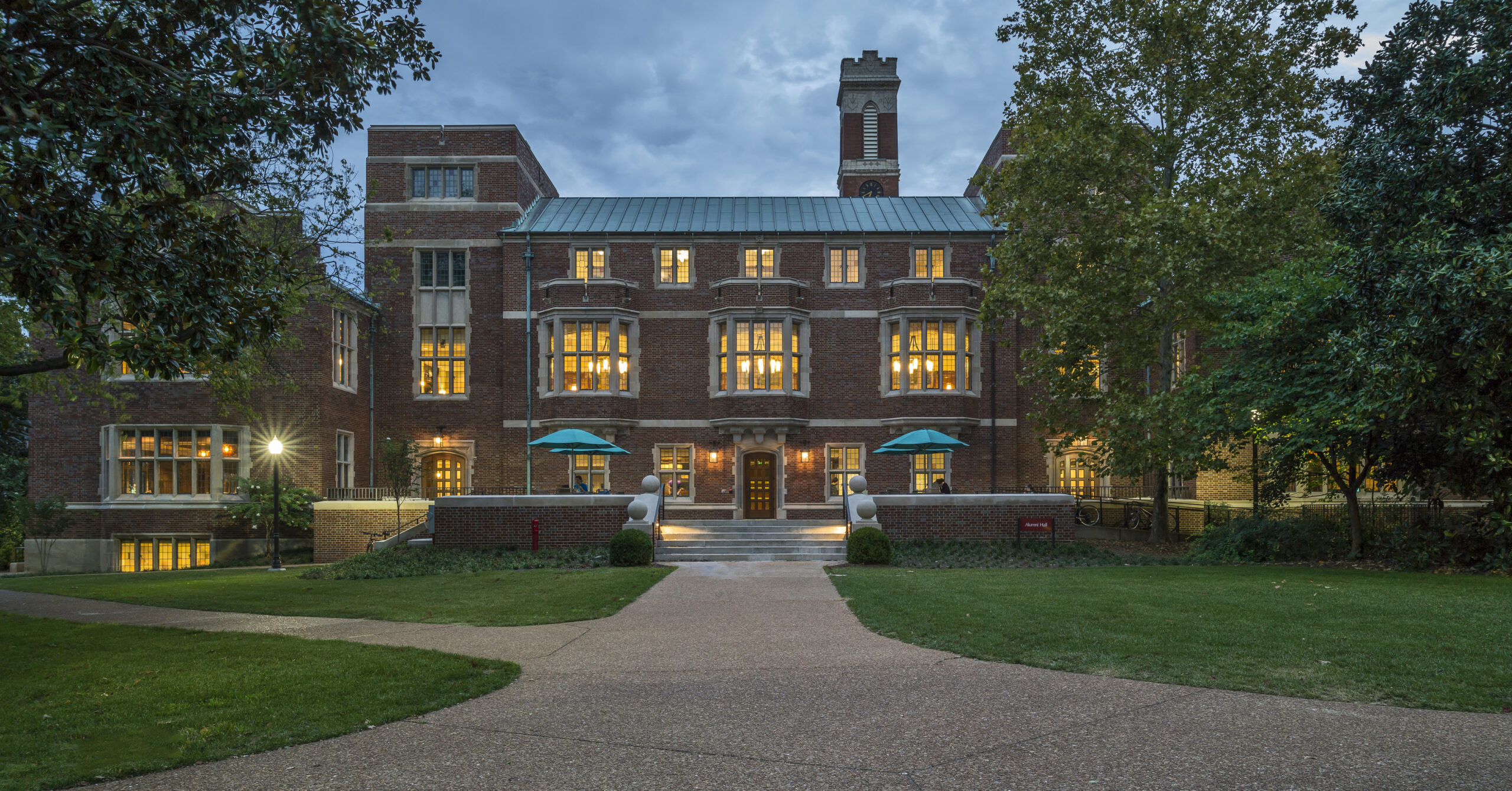 A picture of a stately brick building, sitting at the end of a footpath amid a lawn. The lighting is dusky, emphasizing the lights on in every window.