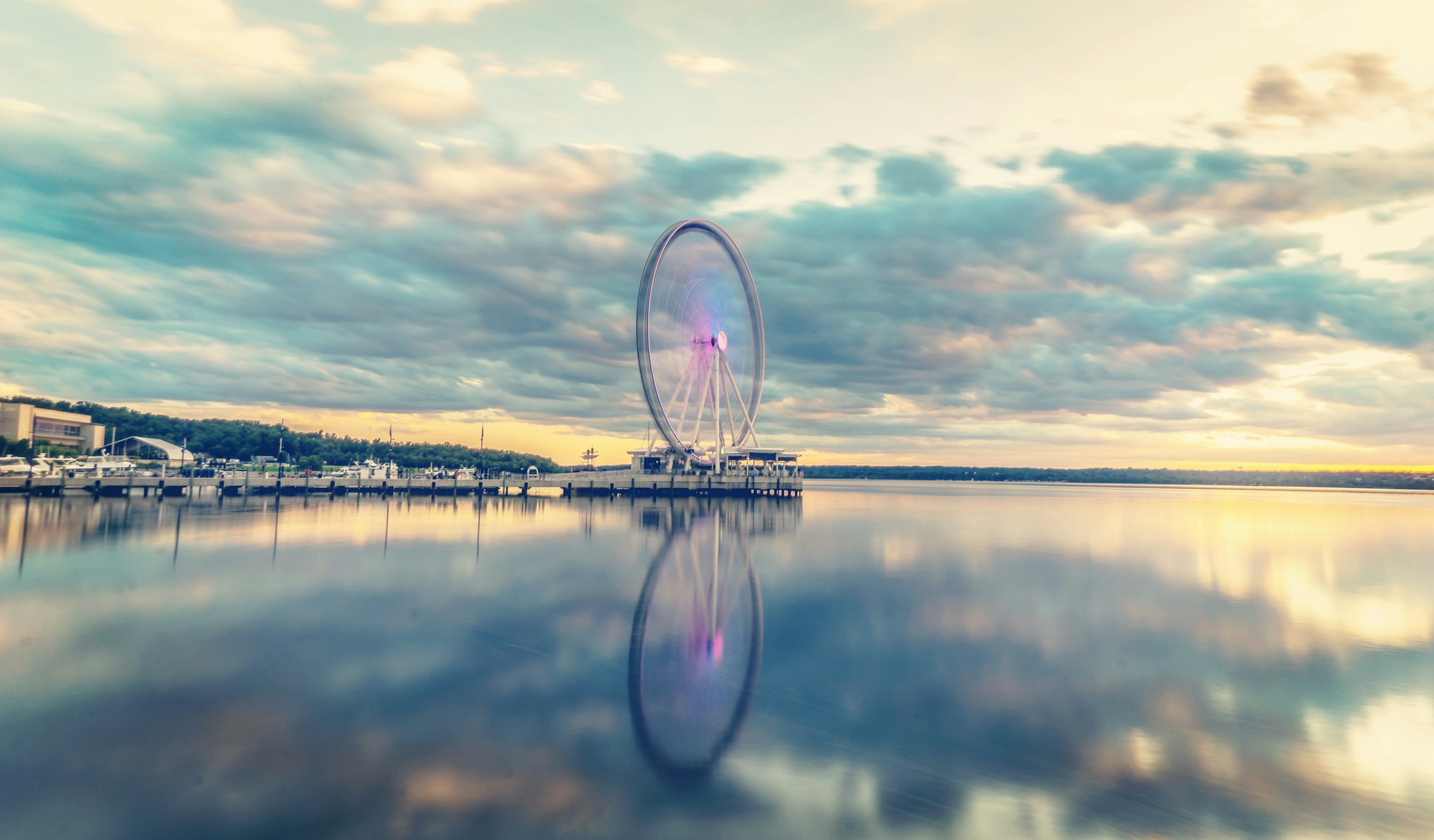 Brightly lit ferris wheel sitting on the horizon in front of National Harbor, Maryland. The ferris wheel is reflected in the light of the Potomac river.