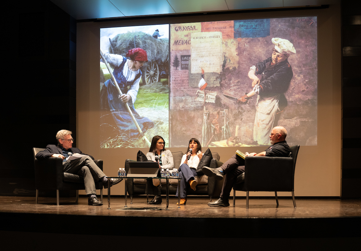 Four people sit on a stage engaged in a panel discussion, with two paintings projected against a backdrop.