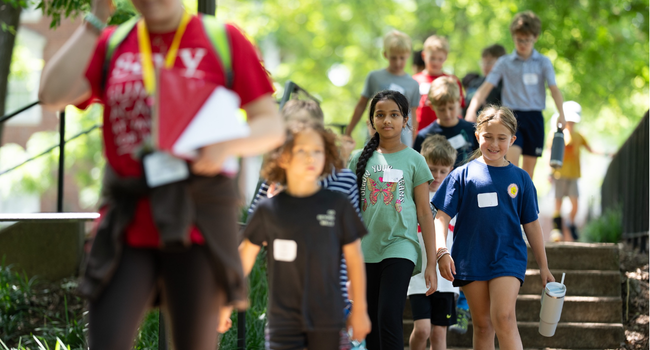 Group of children wearing name tags walk down outdoor steps on a leafy campus, led by an adult counselor.