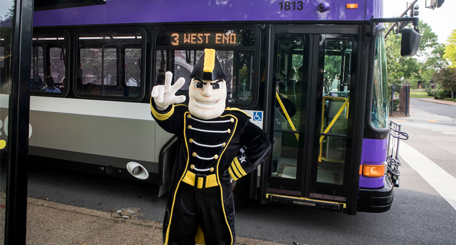 Vanderbilt University mascot dressed in a black and gold uniform stands in front of a purple and white bus displaying the route '3 West End.' The scene features a sidewalk, trees, and part of a historic brick building in the background.