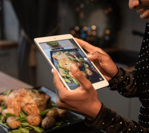 Pictured: Hands hold an electronic device to snap a photo of a homemade turkey. This image represents the concept of "phone eats first" - a trend where people take photos of their food before eating it.