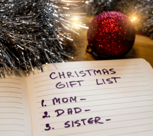 Pictured: A festive spread with tinsel, a red sparkle ornament, warm lights, and a lined notebook. The notebook is open to a page that says "Christmas Gift List." The list reads mom, dad, and sister.