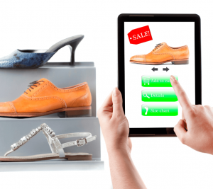 Pictured: Two hands hold a tablet in front of a display of shoes. On the tablet screen is one of the shoes with a sale tag. It is implied that the shopper could save money by buying the shoes online instead of in the store.