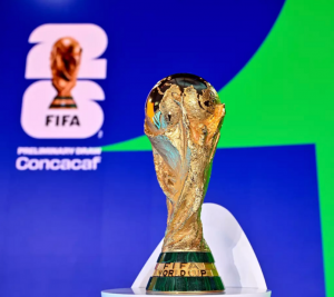 Pictured: the World Cup trophy is in front of a blue and green background. The background also has the 2026 Men's World Cup logo on it.