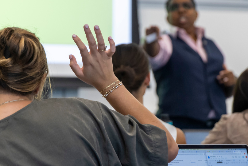 Pictured: Professor Jen Riley encourages student participation in her classes. This image shows a student is raising their hand in Riley's class. Riley is pointing toward the student to indicate it is their turn to speak.