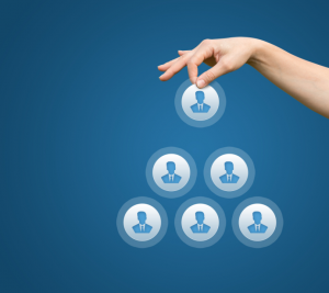 Pictured: On a blue background, a hand is placing an icon of a person at the top of a pyramid. The pyramid represents the hierarchical structure of a workplace.