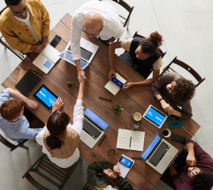 Pictured: A group of 8 people are gathered at a table with laptops. They are working together, and sharing ideas. 