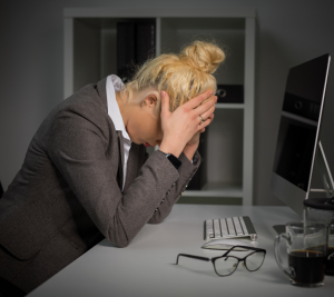 Pictured: A woman dressed in business-attire is sitting at a desk cradling her head in her hands. The image is intended to represent a neurodivergent professional struggling to confirm to a workplace designed by neurotypical standards.