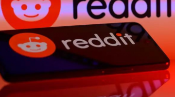 Pictured: The Reddit logo is in the background on the image. A phone is in the focus of the image with the reflection of the Reddit logo on the screen.