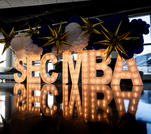 Pictured: In marquee letters that are white, SEC MBA is spelled out. Above the letters, a balloon display hangs from a ceiling.