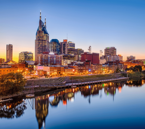Pictured: A photo taken over the Cumberland River, the Nashville city skyline is shown in the evening. The buildings are reflecting off the water and the sun is setting.
