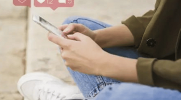 Pictured: A person is sitting with their legs crossed and their hands in their lap. They are holding a phone. Above the phone are graphics with notifications indicating likes and comments on social media.