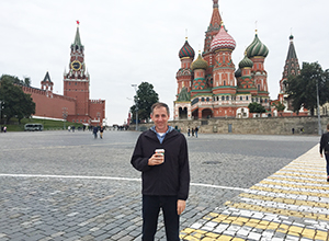 Mark Pettus holding a cup of coffee and standing on pavement in Moscow with a clock tower and colorfully painted, domed building behind him
