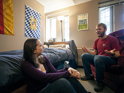 a student sitting on the floor and leaning against a bed talks with a student sitting in an upholstered chair in a dorm room with German and Bavarian flags on the wall