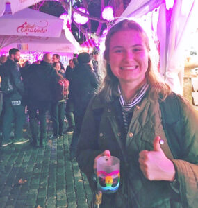 Jacquelyn Olson gives a thumbs-up sign and holds a mug of hot drink while standing outdoors in front of a tent at nighttime in a town square