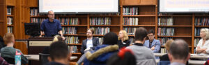 one faculty member stands at a lectern and four others sit at a long table in front of a book-lined room of seated students and faculty