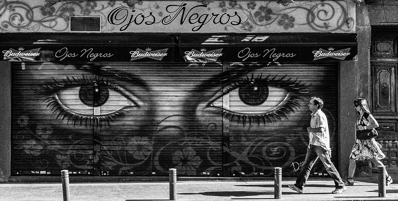 A streetside wall with eyes painted on it and two people walking by.