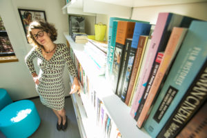 A headshot Jennifer Fay, a white woman with curly brown hair wearing glasses and a patterned dress standing in her office with her hand on her hip