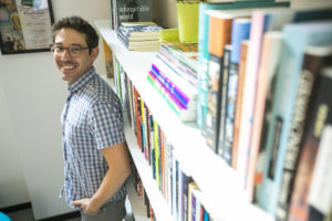 A headshot of Jonathan Rattner, a white man with dark hair and stubble wearing glasses and a plaid button down standing against a bookshelf and looking over his shoulder at the camera