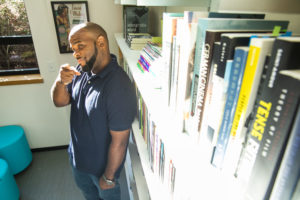 A headshot of Jonathan Waters, a black man with a shaved head and short facial hair wearing a navy polo standing next to a bookshelf and pointing at the camera