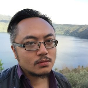 A headshot of Iggy Cortez, an Asian man with pushed back dark hair and dark facial hair and glasses with a lake in the background