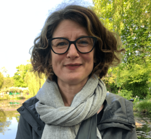 A headshot of Jennifer Fay, a white woman with curly brown hair wearing glasses and a grey scarf with trees and water in the background