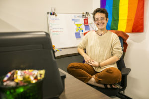 Cassidy Parkison, a white woman with short brown hair wearing a beige sweater and sitting cross-legged in an office chair with a pride flag behind her